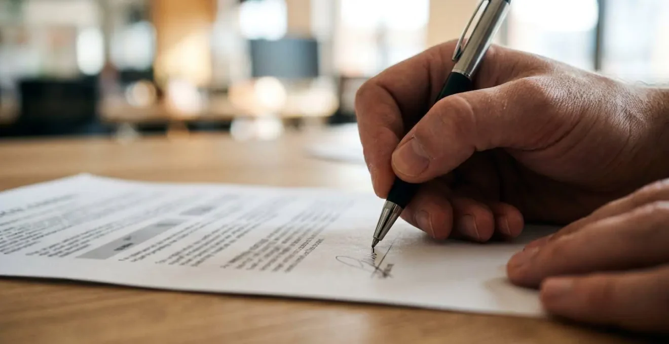 Gros plan sur une main signant un document officiel avec un stylo de qualité sur un bureau en bois clair, arrière-plan flouté